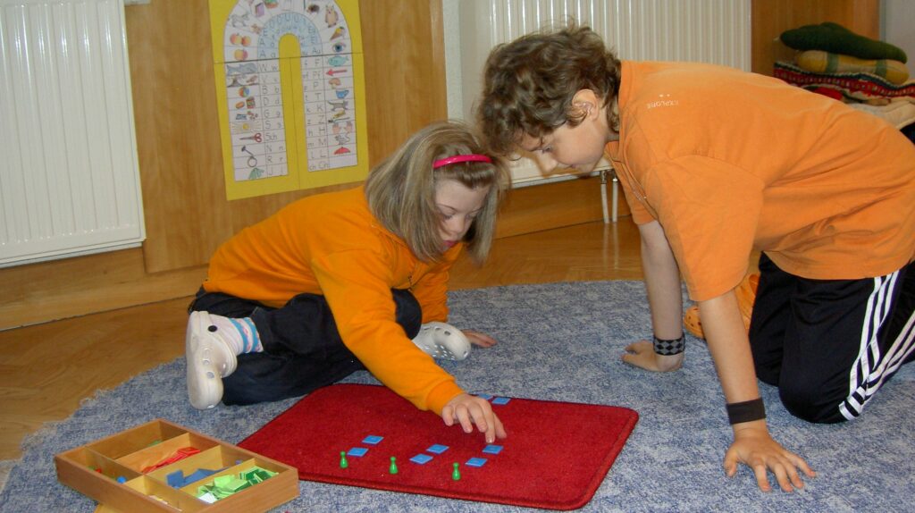 Das Bild zeigt ein Mädchen mit Down Syndrom arbeitet gemeinsam mit einem anderen Schüler an einer Division - mit Montessorimaterial
