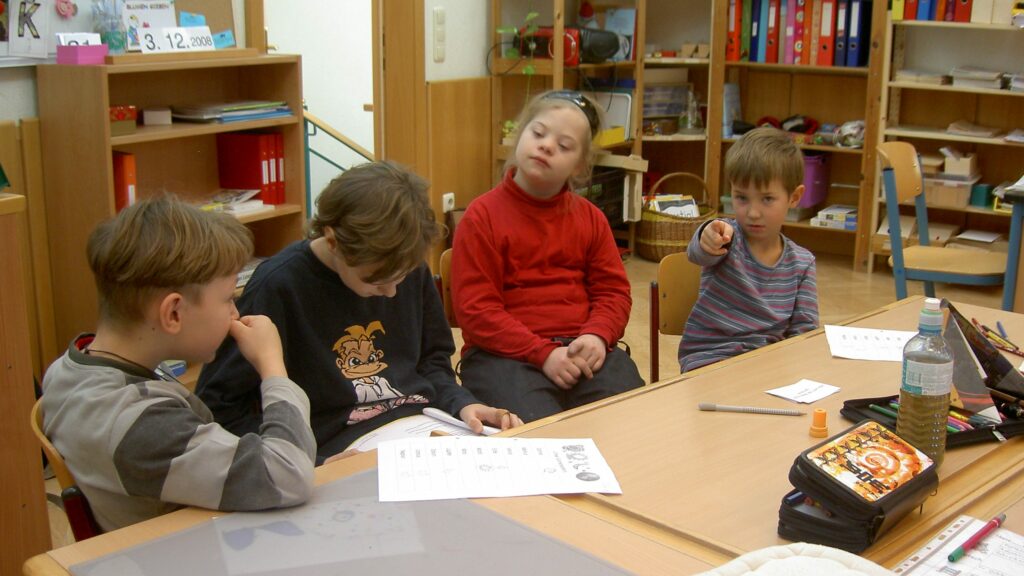 Das Bild zeigt vier Kinder, die gemeinsam an einem Tisch sitzen. Eines der Kinder ist ein Mädchen mit Down-Syndrom. Die anderen Kinder hören aufmerksam zu während der "Experte" aus seinen Notizen vorträgt.